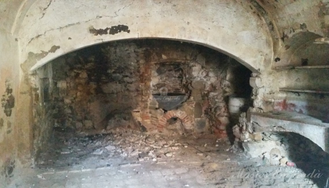 Vista de la cocina con el hogar y el horno bajo cubierta de la chimenea a la izquierda de la imagen y dos vasijas de cenizas para blanquear la ropa