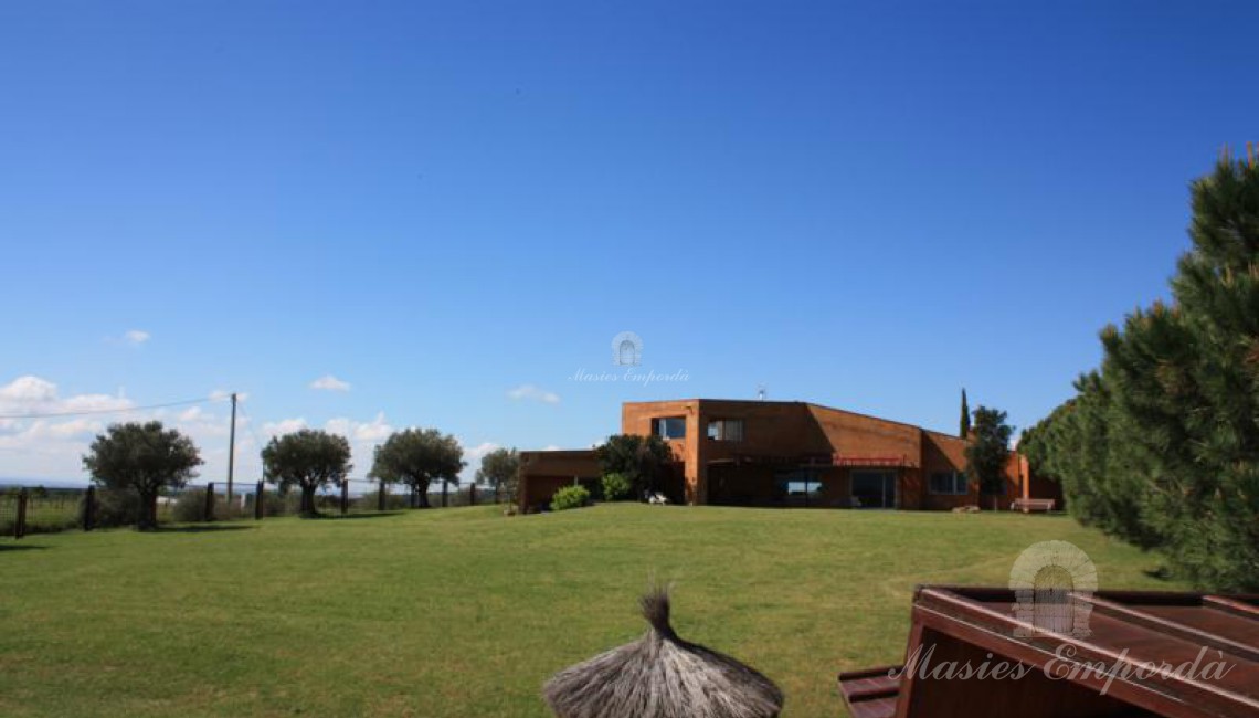 Vista del frontal de la casa desde la zona de la piscina en el jardín con unos olivos centenario que se perfila en un cielo azul típico del Alt Empordà 