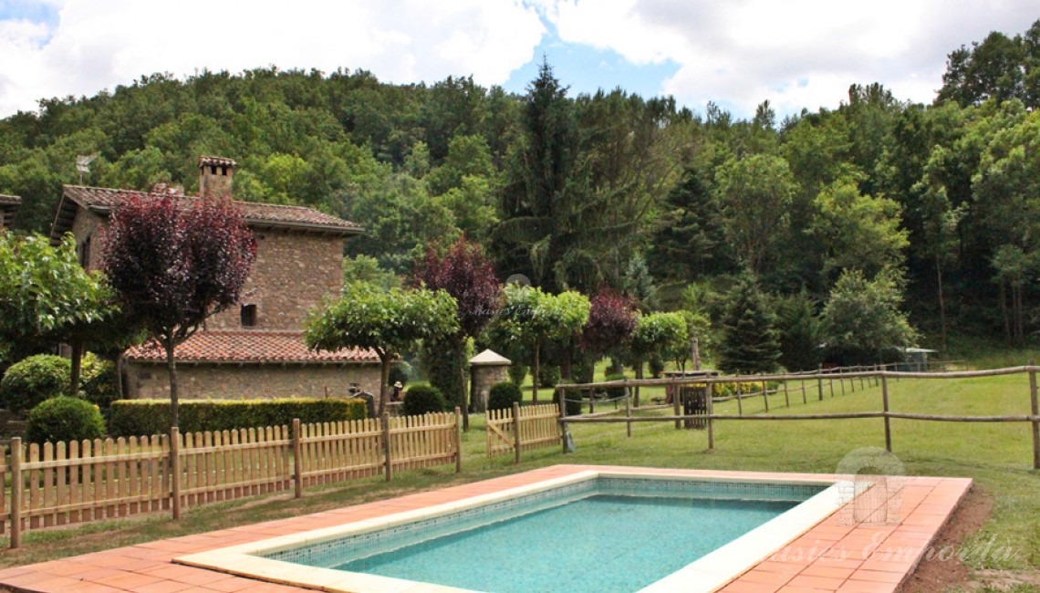 Detalle de la piscina con la masía al fondo y rodeada del jardín 